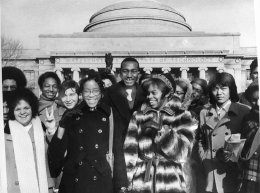 scan of black and white photo with over half a dozen students both men and women, wearing coats in the foreground with the 77 Mass Ave MIT entrance in the background.